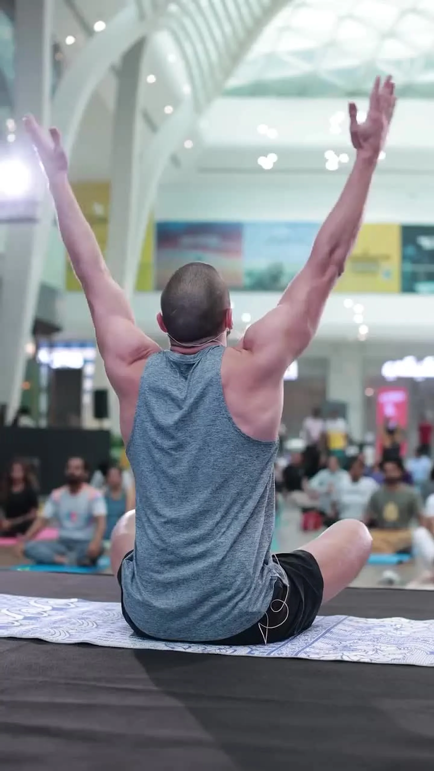 A man leading a seated yoga session, arms raised overhead, in a mall atrium with a class practising behind him